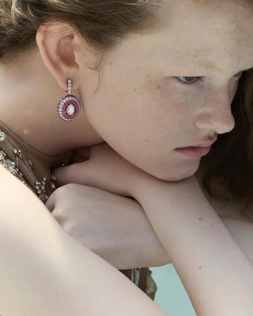 Close-up of a fair-skinned woman wearing round, ornate drop earrings with pink and white stones, showcasing jewelry.