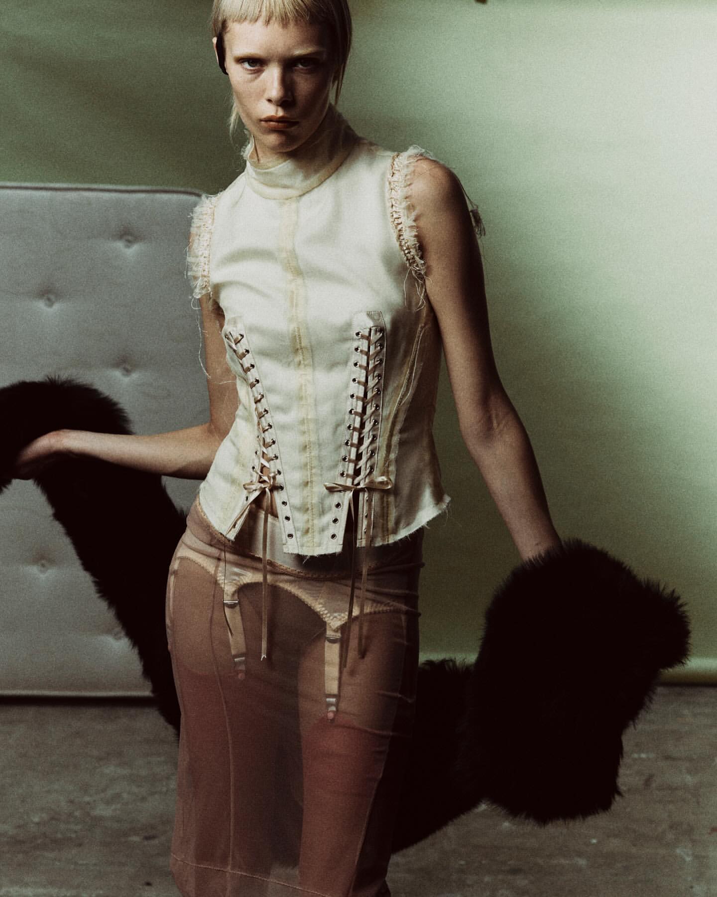 A fashion shot of a slender woman in a corset top and sheer skirt holding a fur stole against a pale green backdrop in a studio setting.
