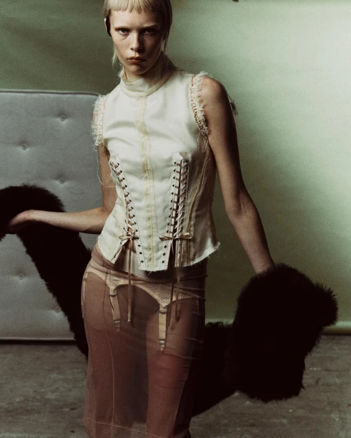 A fashion shot of a slender woman in a corset top and sheer skirt holding a fur stole against a pale green backdrop in a studio setting.