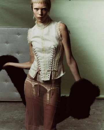 A fashion shot of a slender woman in a corset top and sheer skirt holding a fur stole against a pale green backdrop in a studio setting.