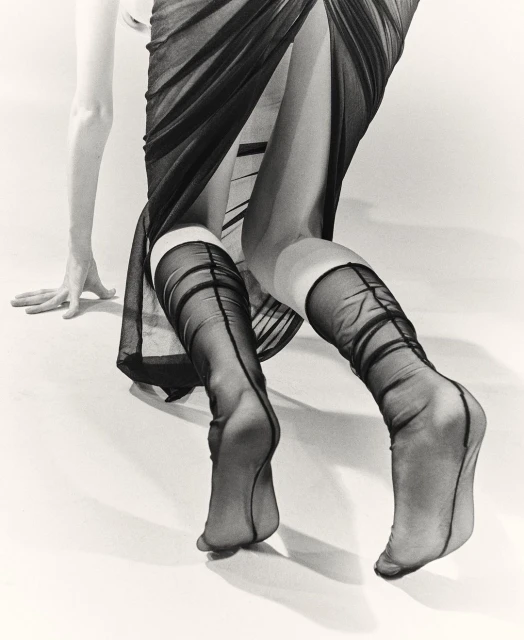 A fashion shot featuring the lower half of a person wearing black, patterned stockings and a sheer dress against a light background.