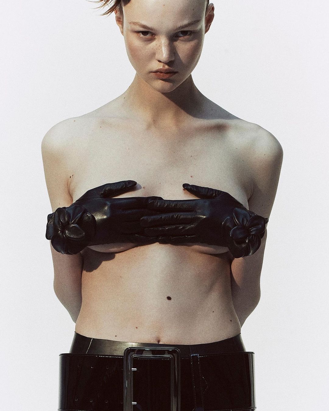 Model with short hair wearing black leather gloves and a patent belt against a plain white background by Melanie and Ramon.