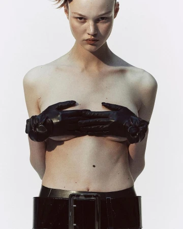 Model with short hair wearing black leather gloves and a patent belt against a plain white background by Melanie and Ramon.