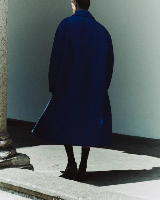 Person in a dark blue overcoat viewed from the back, with dark socks and shoes, standing on a stone surface, against a plain wall.