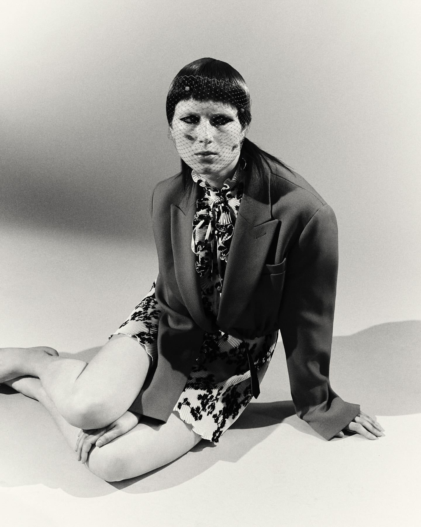 A stylish woman in a blazer and patterned dress poses indoors against a neutral backdrop, wearing a net veil, creating a retro-chic aesthetic.