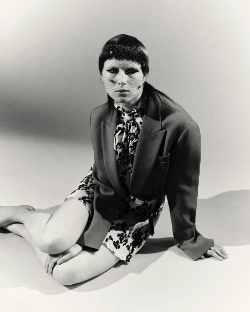 A stylish woman in a blazer and patterned dress poses indoors against a neutral backdrop, wearing a net veil, creating a retro-chic aesthetic.