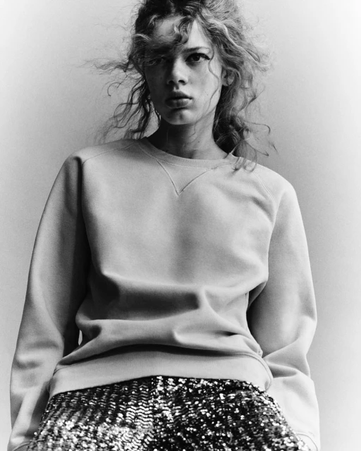 Monochrome portrait of a young woman with wavy hair wearing a sweatshirt and sequined shorts against a neutral studio backdrop.