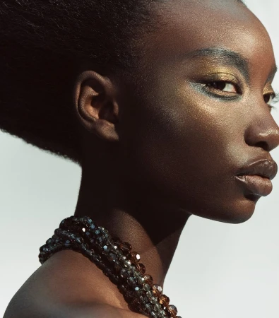 Close-up profile of a black woman with metallic eyeshadow and beaded necklace against a white background