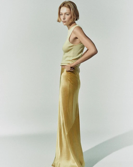 Adult female posing in a studio wearing a light-yellow knit top and floor-length gold silk skirt, creating a fashion-editorial aesthetic.
