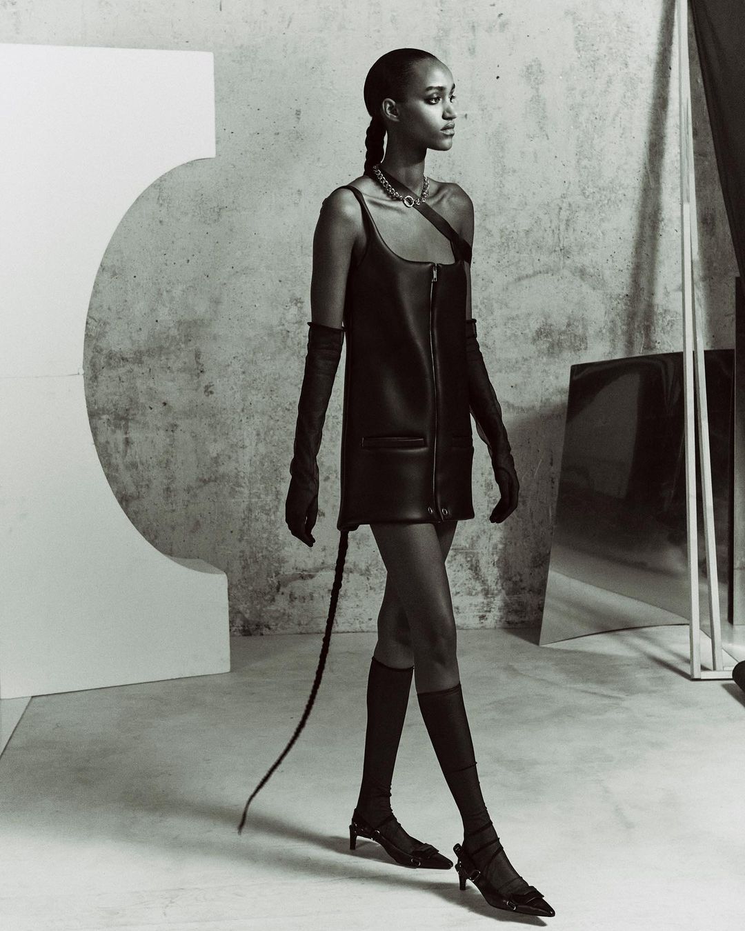 A slender adult female with a braided hairstyle, wearing a black leather dress, long gloves, and low heels, walks in a minimalist photo studio.