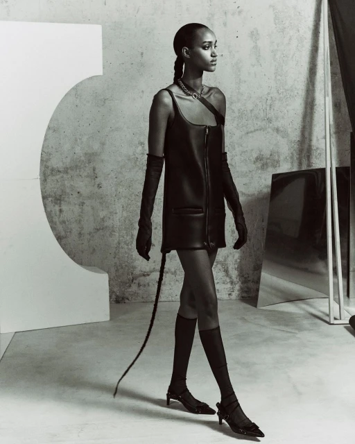 A slender adult female with a braided hairstyle, wearing a black leather dress, long gloves, and low heels, walks in a minimalist photo studio.
