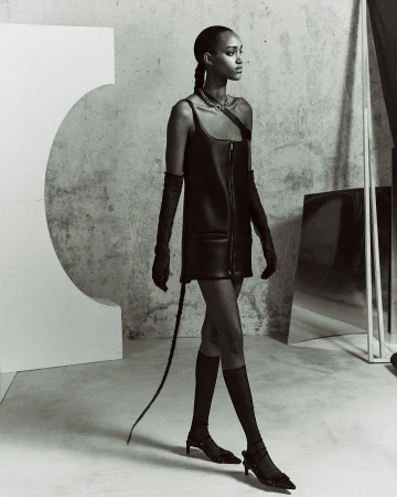 A slender adult female with a braided hairstyle, wearing a black leather dress, long gloves, and low heels, walks in a minimalist photo studio.
