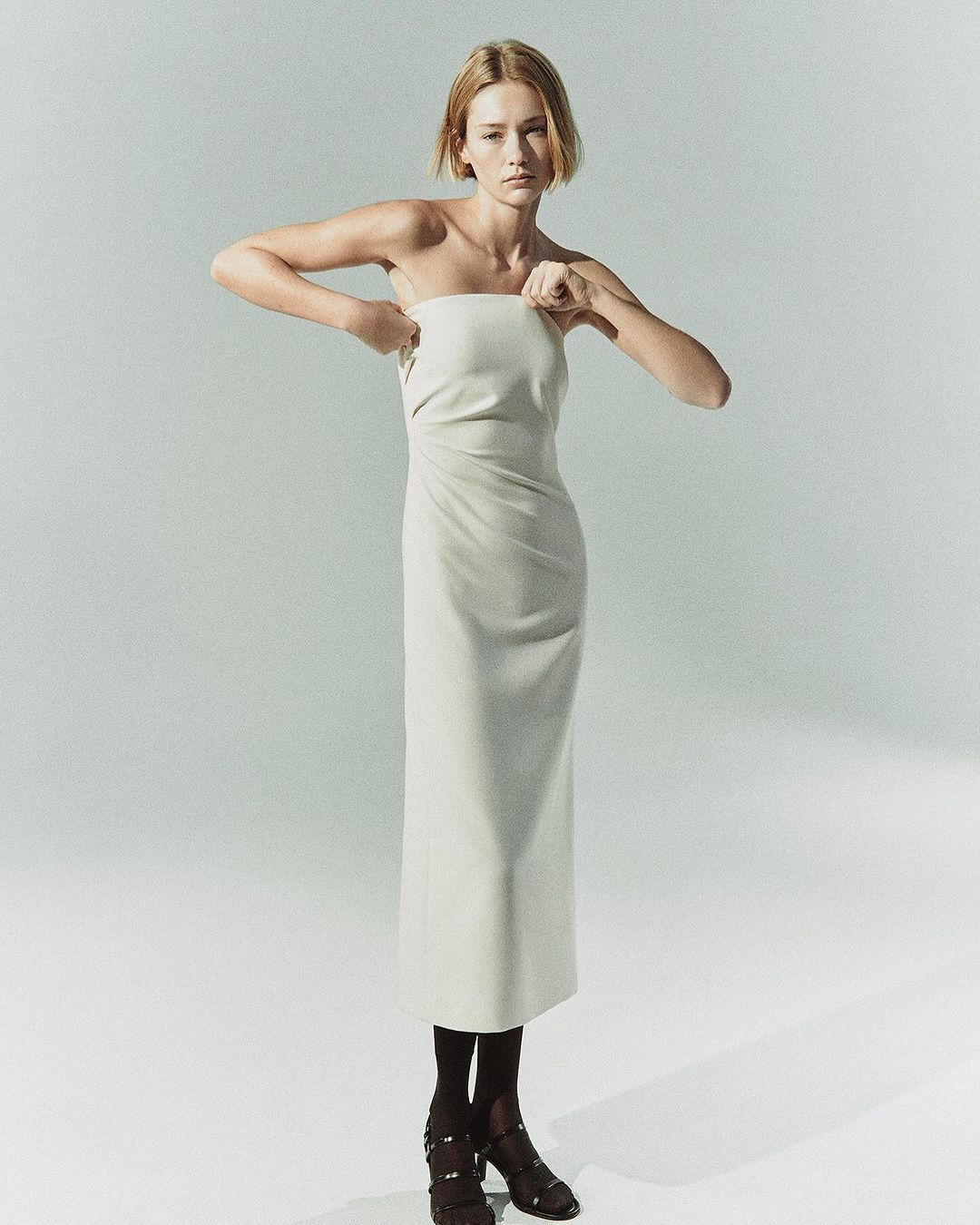 Model in strapless cream midi dress adjusting neckline against a plain pale grey background.