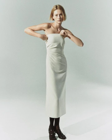 Model in strapless cream midi dress adjusting neckline against a plain pale grey background.