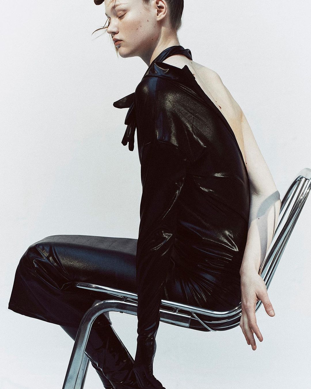 Woman in black vinyl dress seated on a chrome chair in a studio, posing with hands visible, against a neutral white backdrop.