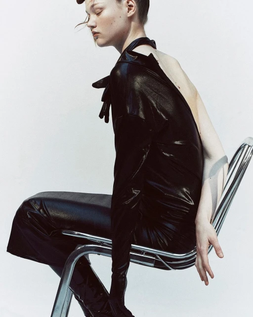Woman in black vinyl dress seated on a chrome chair in a studio, posing with hands visible, against a neutral white backdrop.