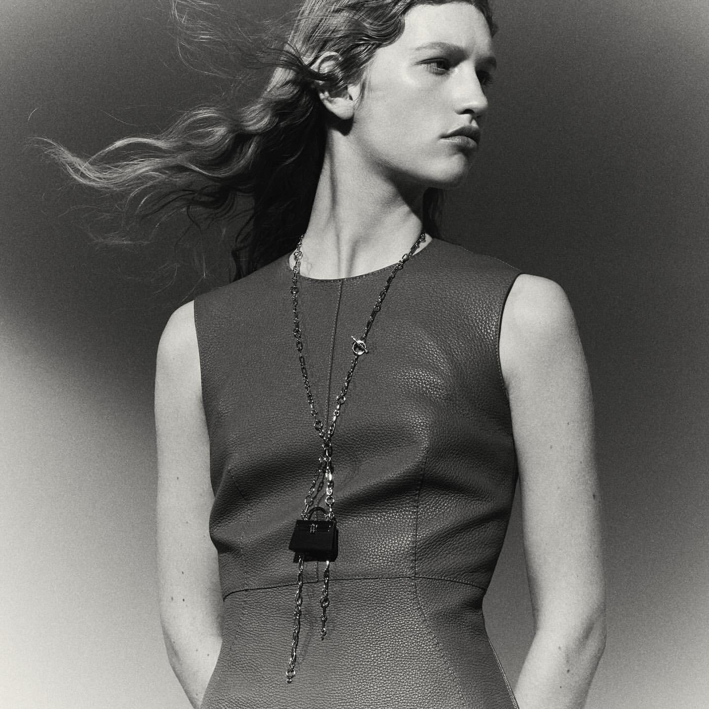 Model in a textured sleeveless dress wearing a long chain necklace with a mini handbag pendant against a plain gradient backdrop