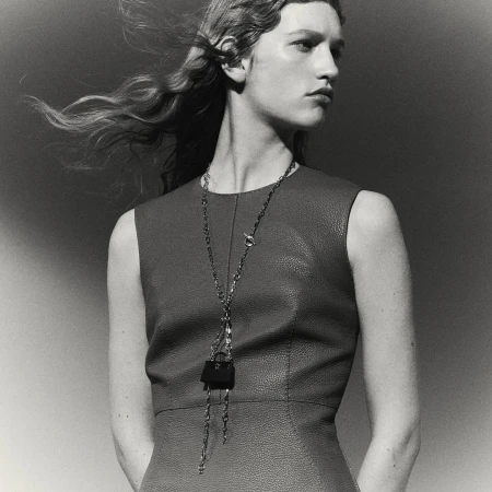 Model in a textured sleeveless dress wearing a long chain necklace with a mini handbag pendant against a plain gradient backdrop