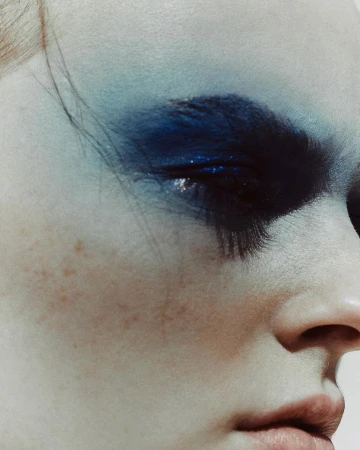 Close-up portrait of a woman with dramatic blue eyeshadow, dark feathered eyelashes, and freckles across her cheek.