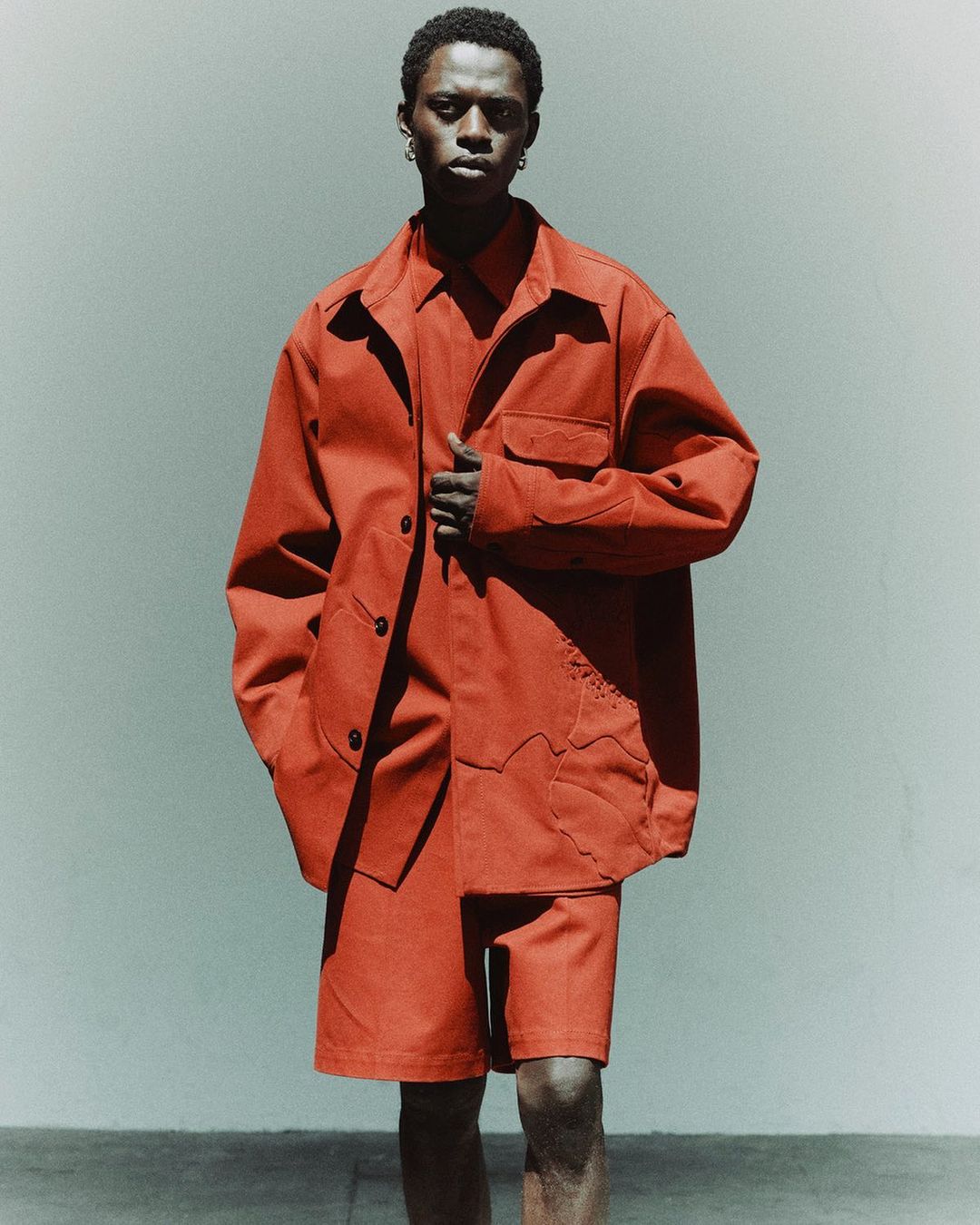 Young Black man in reddish-brown matching collared shirt, shorts, and jacket stands in a photo studio with a neutral background.