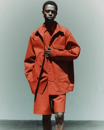 Young Black man in reddish-brown matching collared shirt, shorts, and jacket stands in a photo studio with a neutral background.