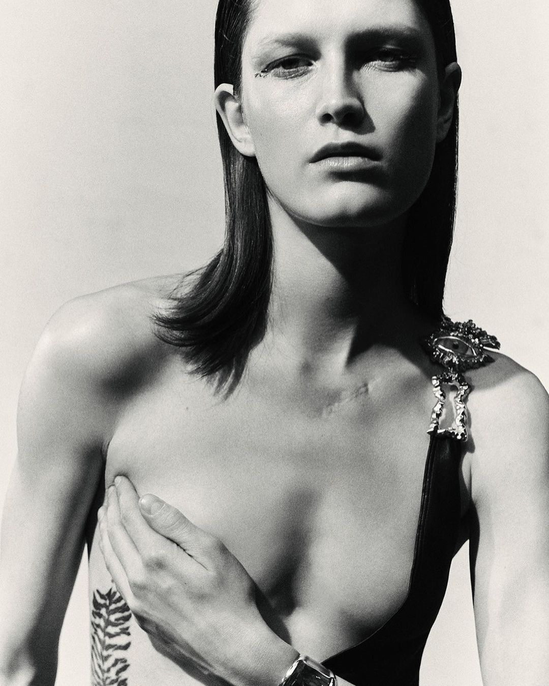 Monochrome fashion portrait of a woman with sleek hair, minimal makeup, and body art, posing in front of a plain, light-colored background.