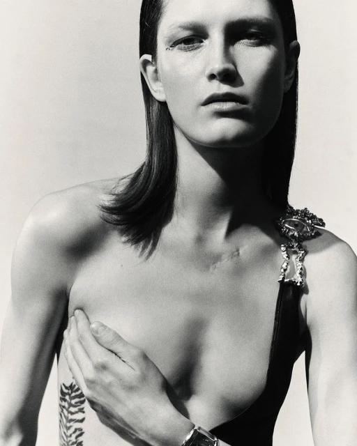 Monochrome fashion portrait of a woman with sleek hair, minimal makeup, and body art, posing in front of a plain, light-colored background.