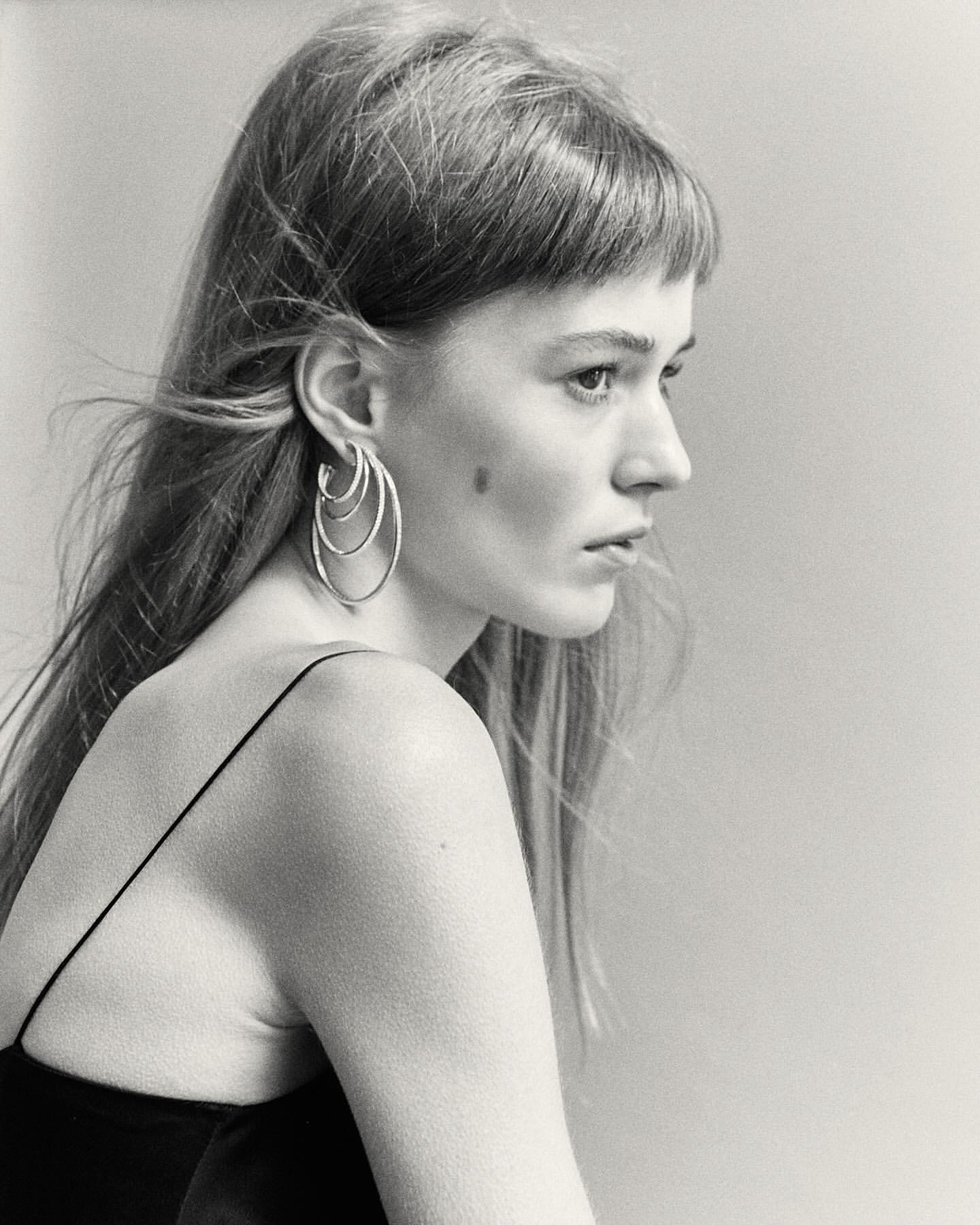 Black and white profile portrait of a woman wearing layered silver hoop earrings and a dark spaghetti-strap top.