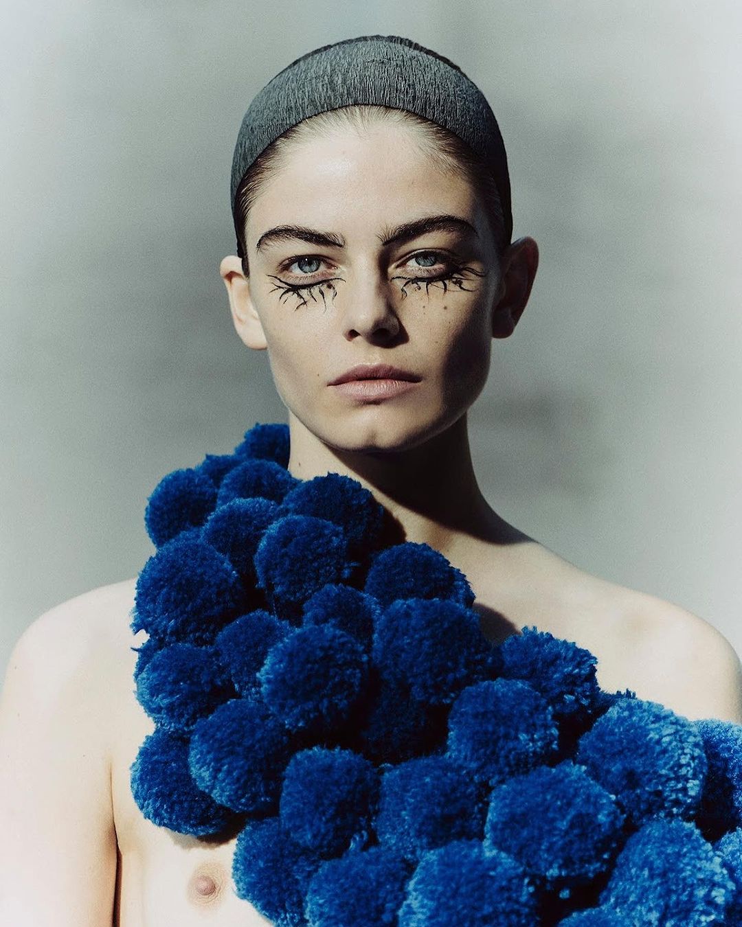 Striking close-up of a bare-chested person with unique eye makeup and a blue pom-pom adornment, against a neutral background.