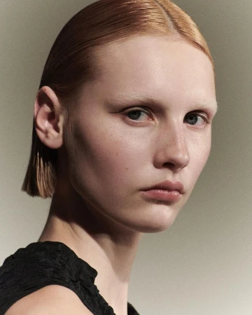 Close-up portrait of a woman with slicked-back ginger hair and pale eyebrows wearing a black top against a grey textured backdrop