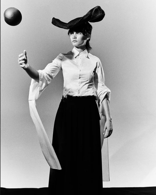 Black and white shot of a woman in a sculptural hat and flowing sleeves, poised near a floating ball, against a white backdrop.
