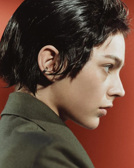Close-up portrait of a young adult woman with short dark hair and a green jacket, facing right against a red backdrop in a studio setting.
