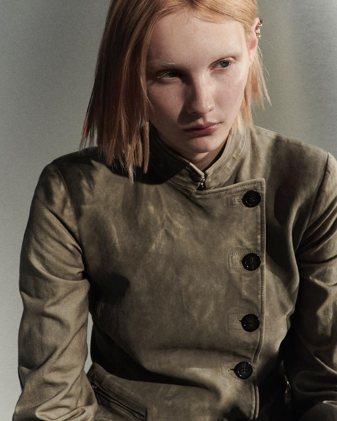 Portrait of a red-haired, fair-skinned woman wearing a buttoned, olive-green jacket against a neutral studio background.