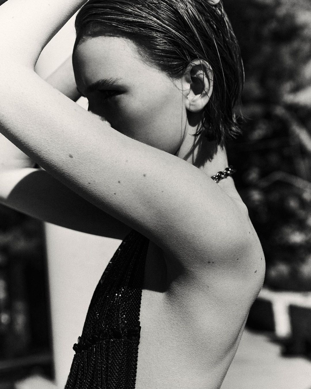 Monochrome portrait of a woman with wet slicked-back hair and minimal makeup, arms raised behind her head, wearing a dark beaded top.