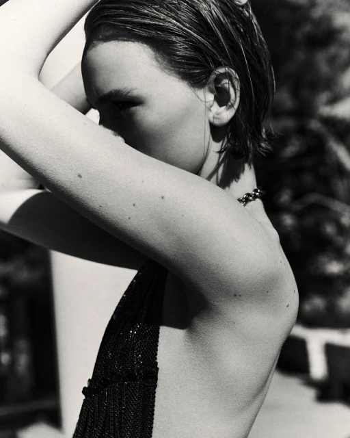 Monochrome portrait of a woman with wet slicked-back hair and minimal makeup, arms raised behind her head, wearing a dark beaded top.