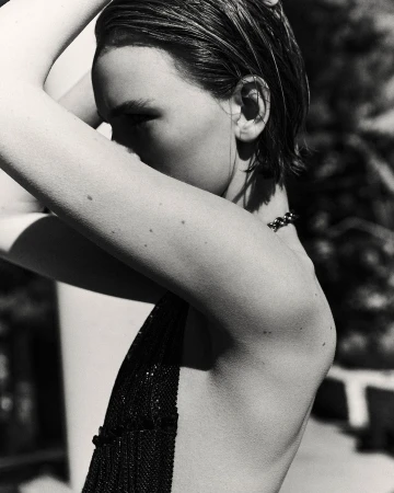 Monochrome portrait of a woman with wet slicked-back hair and minimal makeup, arms raised behind her head, wearing a dark beaded top.