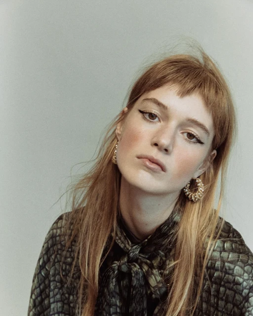 Portrait of a young adult woman with long red hair and heavy black eyeliner wearing a reptile-print blouse against a grey background.