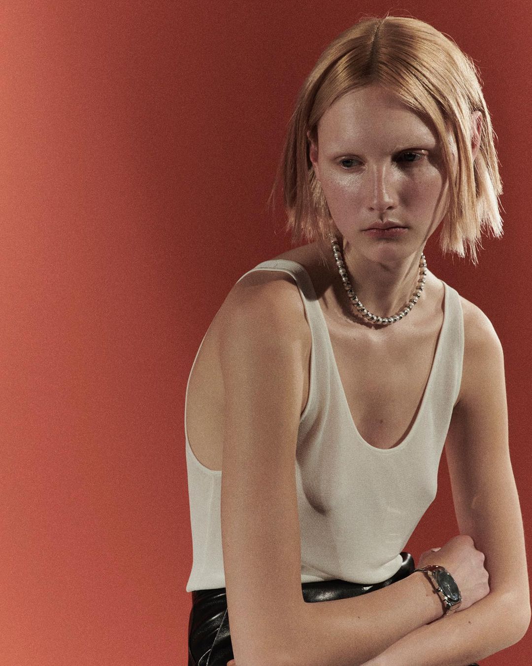 Close-up portrait of a blonde woman in a white tank top and black skirt, wearing a chain necklace, against a reddish-orange background.
