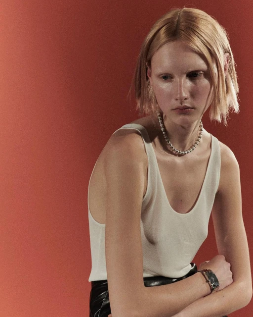 Close-up portrait of a blonde woman in a white tank top and black skirt, wearing a chain necklace, against a reddish-orange background.