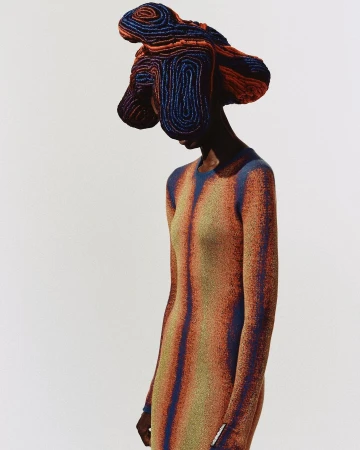 A woman with dark skin wears a copper and blue dress and matching abstract headpiece against a bright white backdrop in a studio shot.