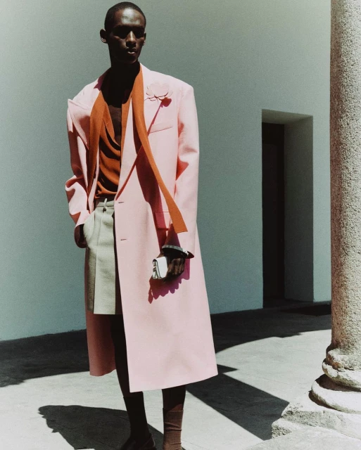 A model poses in a pink overcoat with a light tan sweater and pants outside against a light colored building with a dark doorway.