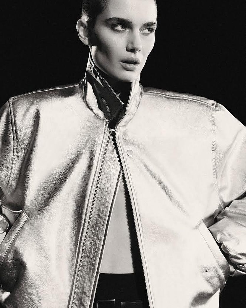 Close-up studio shot of a woman with a shaved head wearing a silver metallic jacket over a black top and trousers, against a black background.