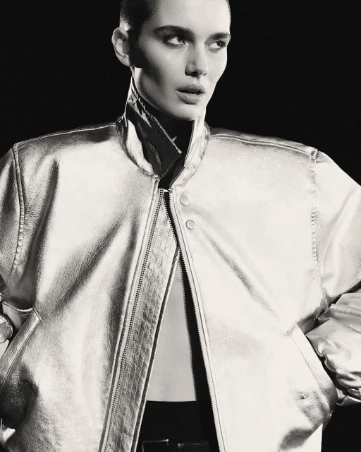 Close-up studio shot of a woman with a shaved head wearing a silver metallic jacket over a black top and trousers, against a black background.
