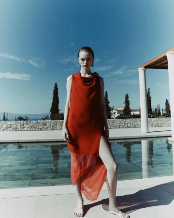 A barefoot woman in a sleeveless red cowl neck dress standing outdoors beside a stone-bordered swimming pool under a blue sky.