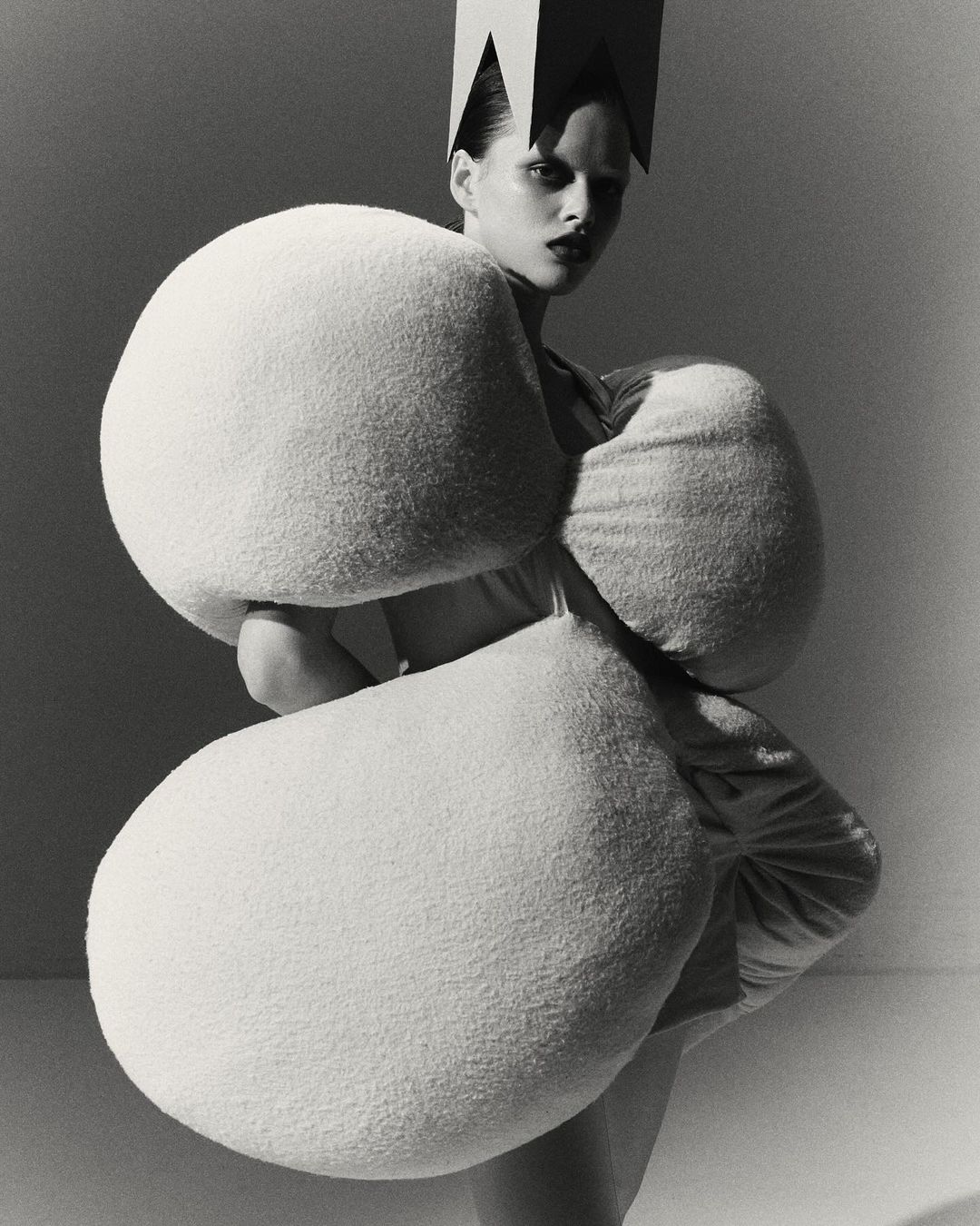 Monochrome studio shot of a stylish adult woman with dramatic makeup wearing an avant-garde crown and sculptural, white padded garment.