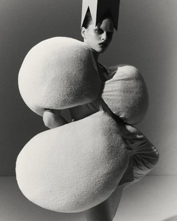 Monochrome studio shot of a stylish adult woman with dramatic makeup wearing an avant-garde crown and sculptural, white padded garment.