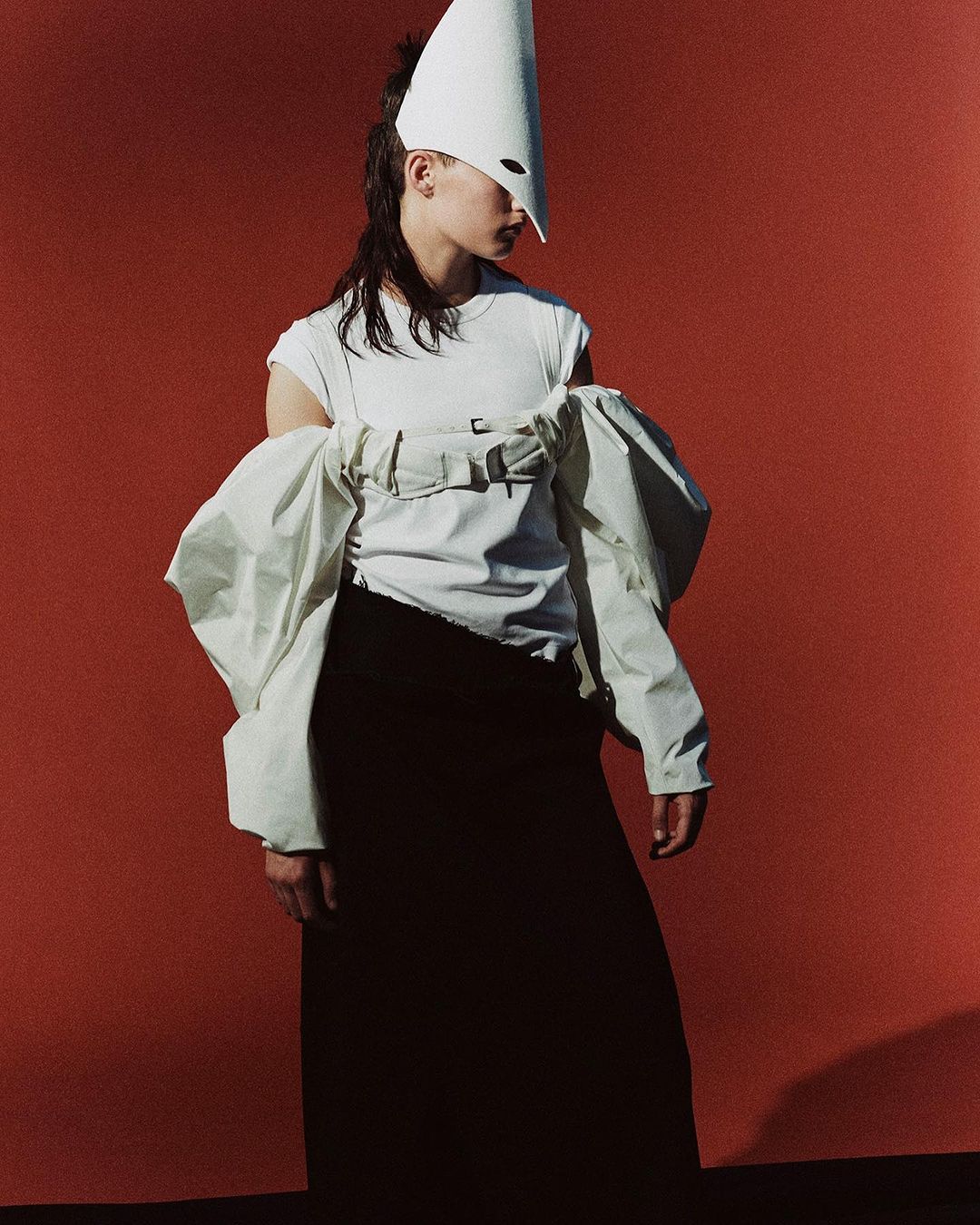 Stylish woman in a white t-shirt and black skirt poses against a solid red backdrop, wearing a white cone hat and oversized sleeves.