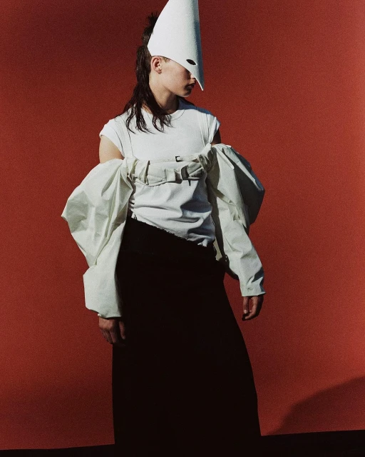 Stylish woman in a white t-shirt and black skirt poses against a solid red backdrop, wearing a white cone hat and oversized sleeves.