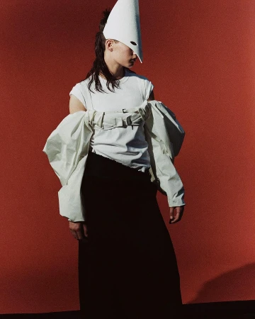 Stylish woman in a white t-shirt and black skirt poses against a solid red backdrop, wearing a white cone hat and oversized sleeves.