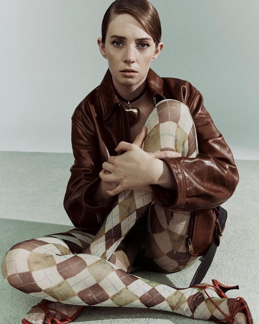 A woman with styled hair sits in a brown leather jacket and patterned tights, gazing calmly at the camera in a studio portrait.